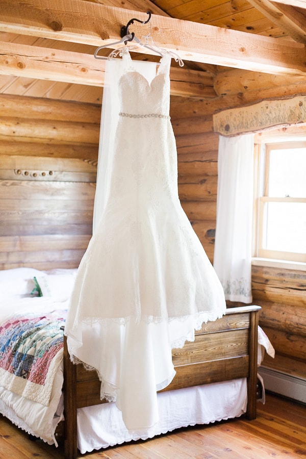 All-over lace wedding gown with illusion straps, sweetheart neckline, and embellished bridal belt. The dress hangs inside a rustic wedding venue in Woodland Park, Colorado. Photo by Rachel Havel; Dress by Danelle's Bridal Boutique
